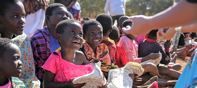 A group of Malawi children sit on the ground holding bags of Nu Skin VitaMeal.
